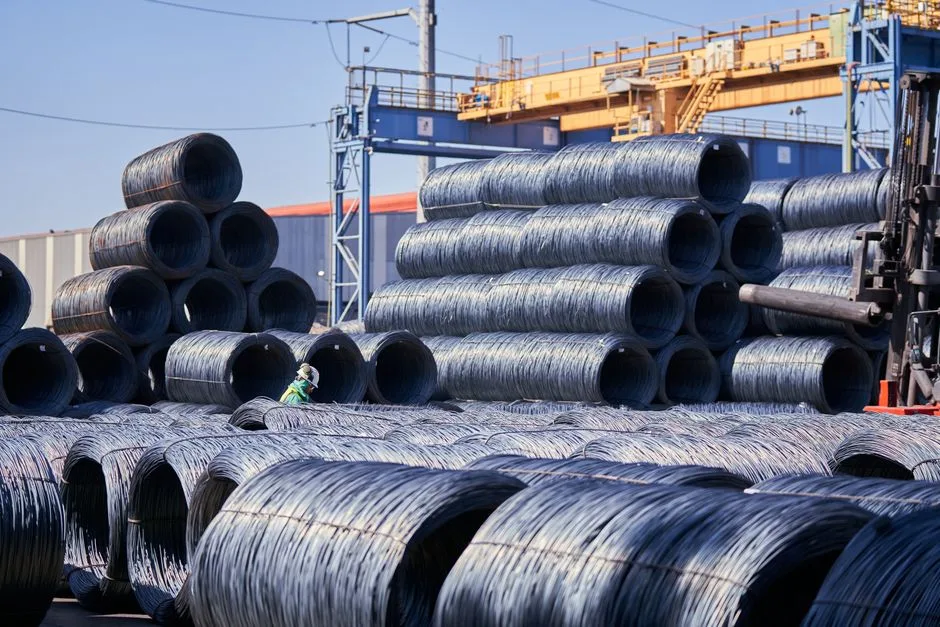 Grandes pilas de bobinas de alambre de acero almacenadas al aire libre en un sitio industrial bajo una brillante luz diurna.