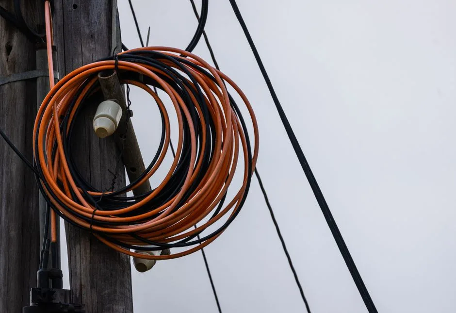 Fotografía en primer plano de cables eléctricos naranjas y negros enrollados en un poste de madera, con un cielo gris de fondo.