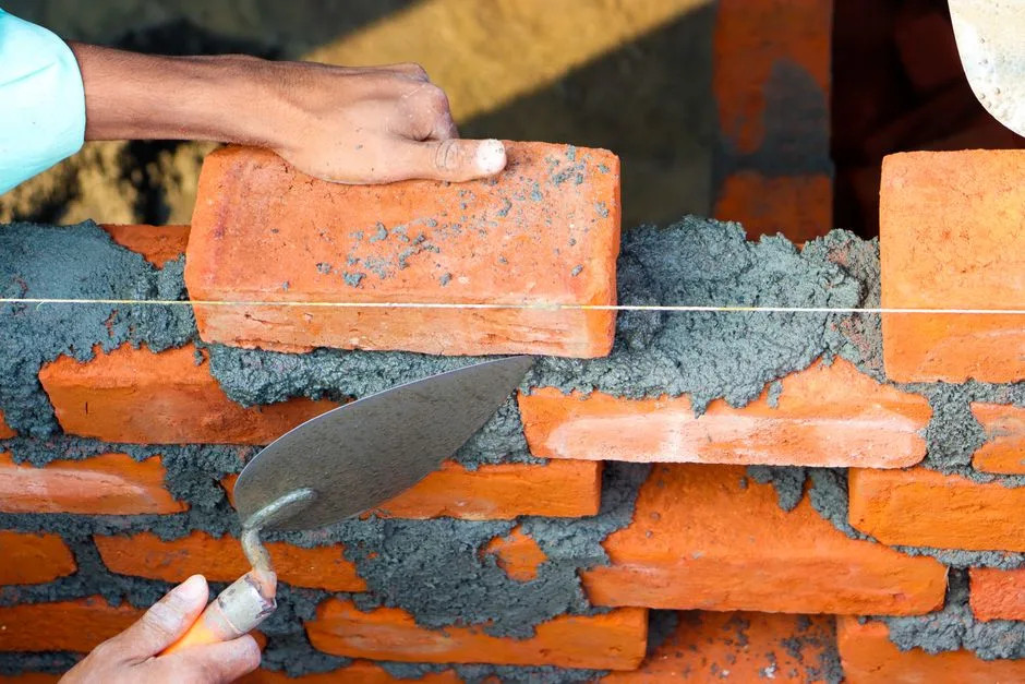 Manos colocando ladrillos con cemento en un entorno de construcción, mostrando un trabajo de albañilería preciso.