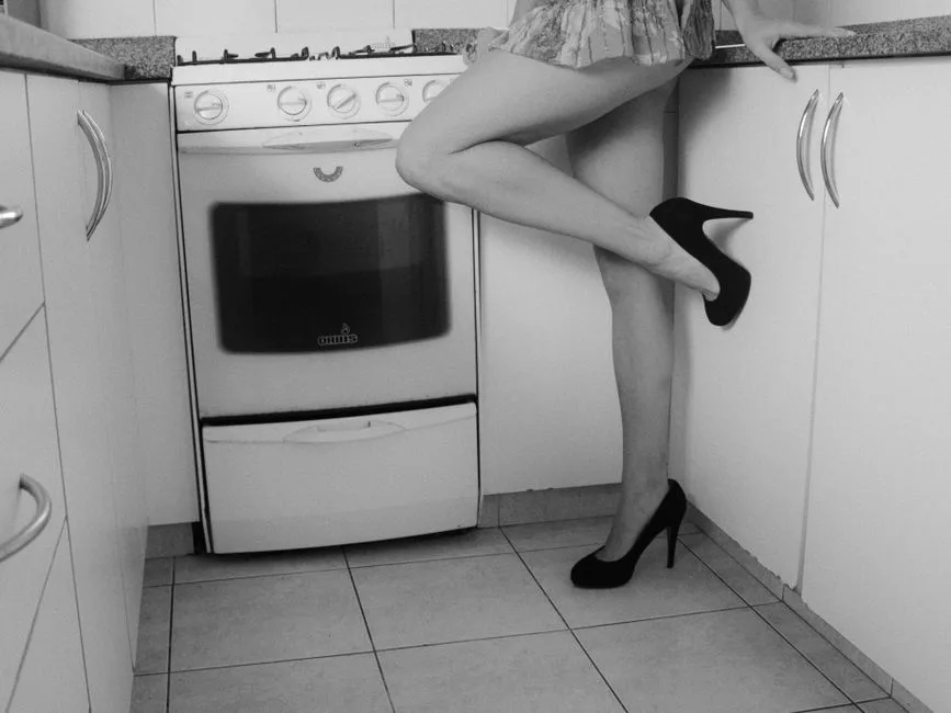 Fotografía en blanco y negro que muestra las piernas de una mujer con tacones altos en un rincón de la cocina.